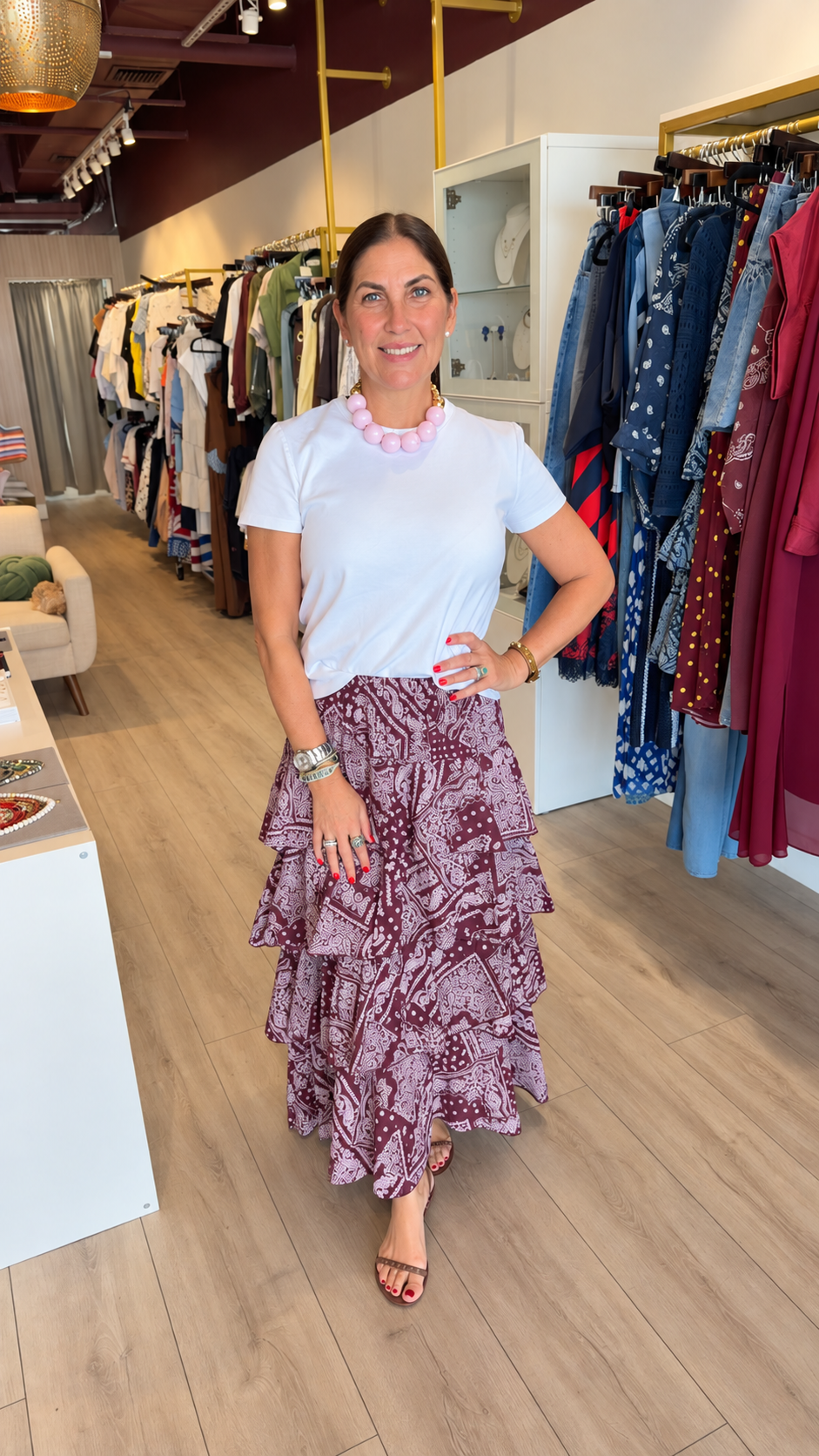 A woman posing in a clothing boutique, wearing a white short-sleeve crewneck top and a long, tiered burgundy and purple patterned maxi skirt, paired with a pink collar necklace.