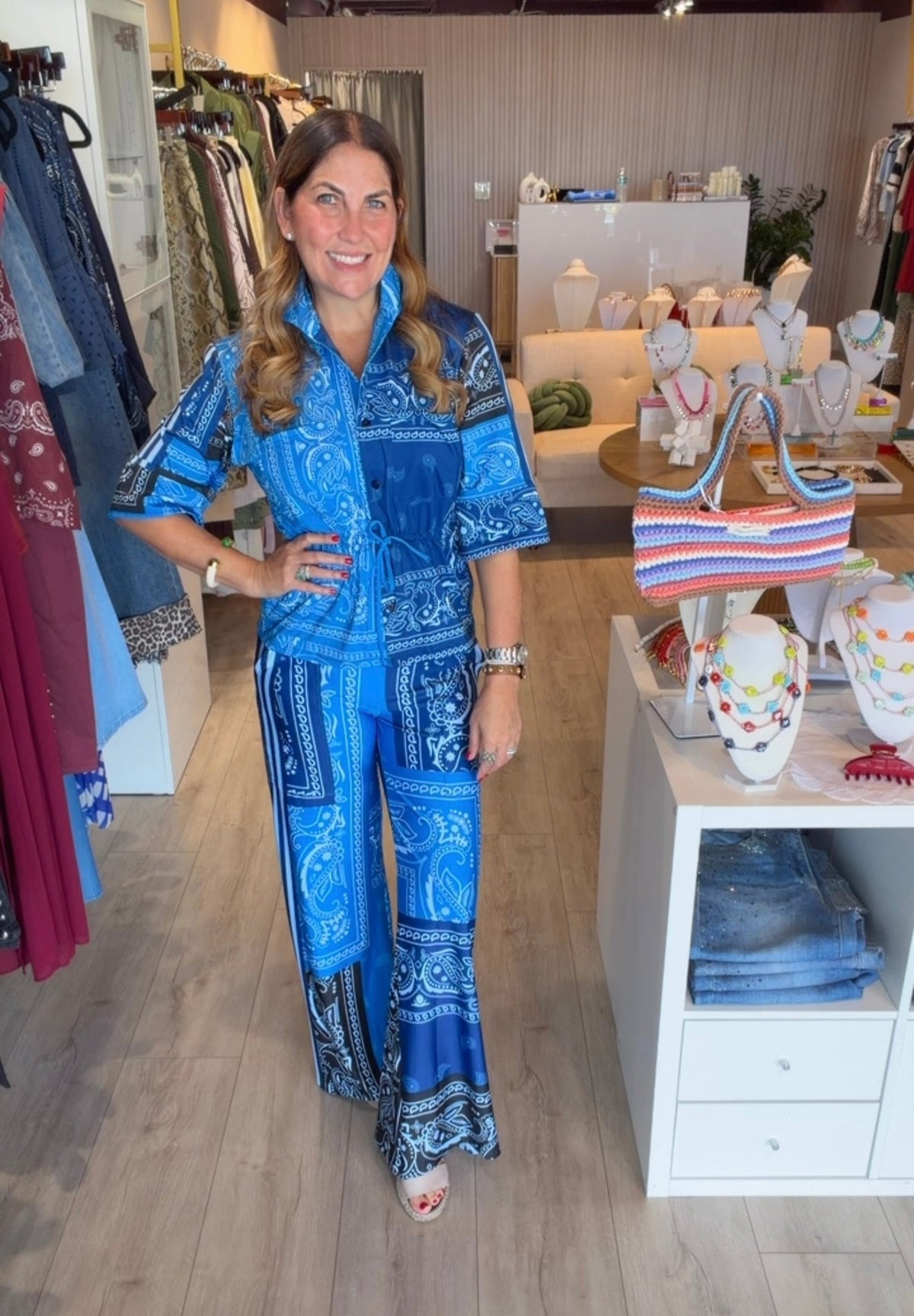 A woman modeling a blue and white paisley print two-piece set consisting of a short-sleeve button-up shirt and matching wide-leg pants, standing inside a boutique store.