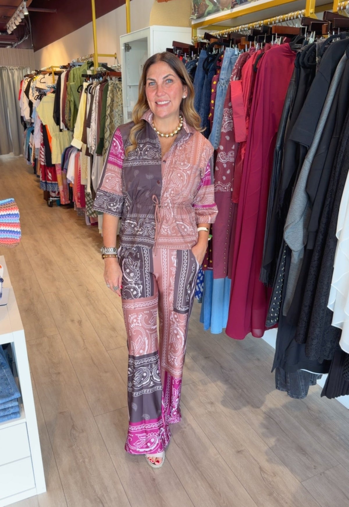 A woman wearing a matching two-piece printed jumpsuit stands inside a clothing boutique. The jumpsuit features a pink, purple, and brown paisley and bandana print pattern.