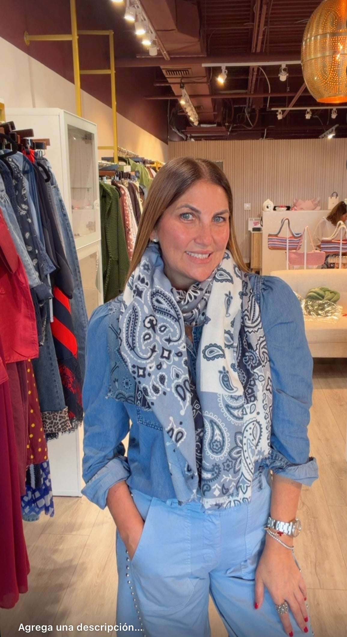A woman wearing a light blue denim shirt and matching light blue pants, accessorized with a blue and white paisley scarf, standing inside a clothing boutique.