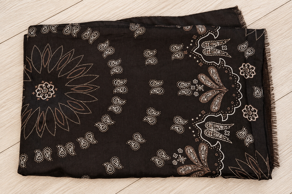 A folded black scarf with intricate beige and white paisley and floral patterns, laid flat on a light wood floor.