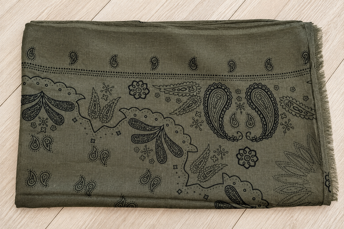 An olive green scarf with a black paisley and floral bandana pattern, folded flat on a light wood floor.