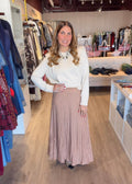 A woman modeling a white long-sleeve knit top and a dusty pink pleated maxi skirt inside a clothing boutique.