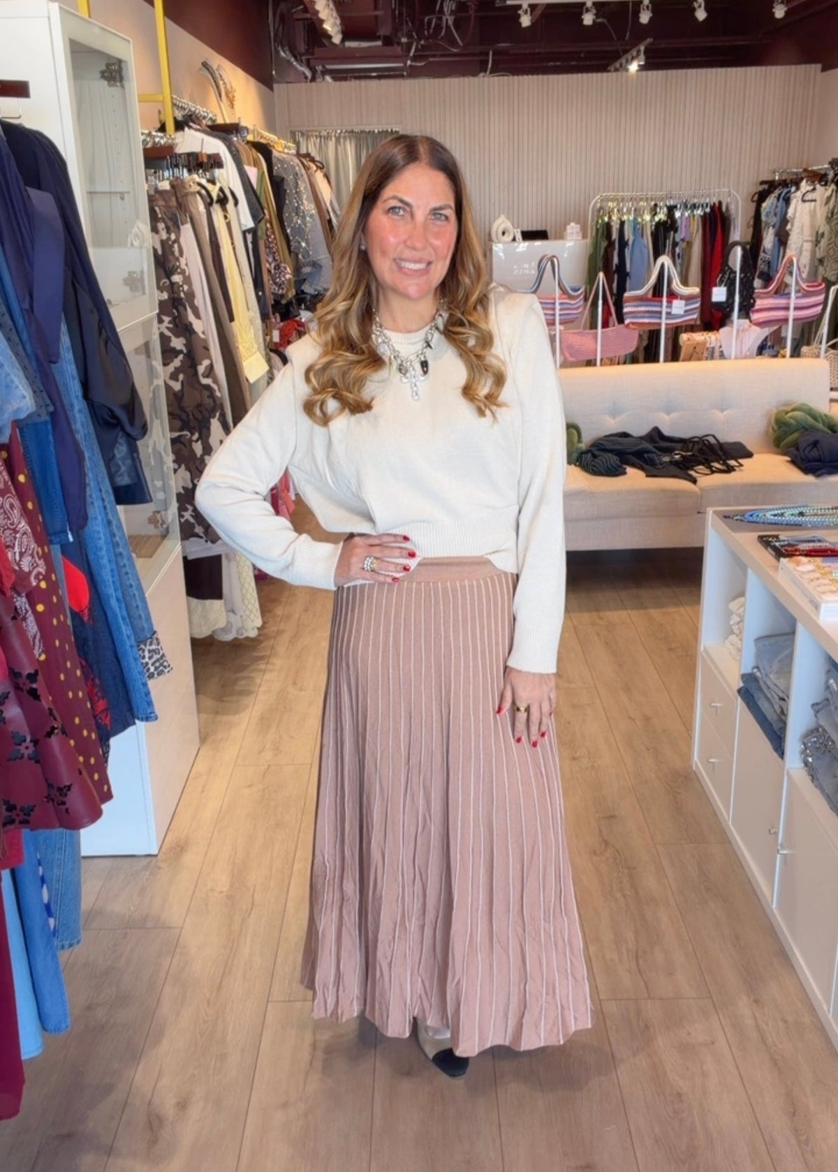 A woman modeling a white long-sleeve knit top and a dusty pink pleated maxi skirt inside a clothing boutique.