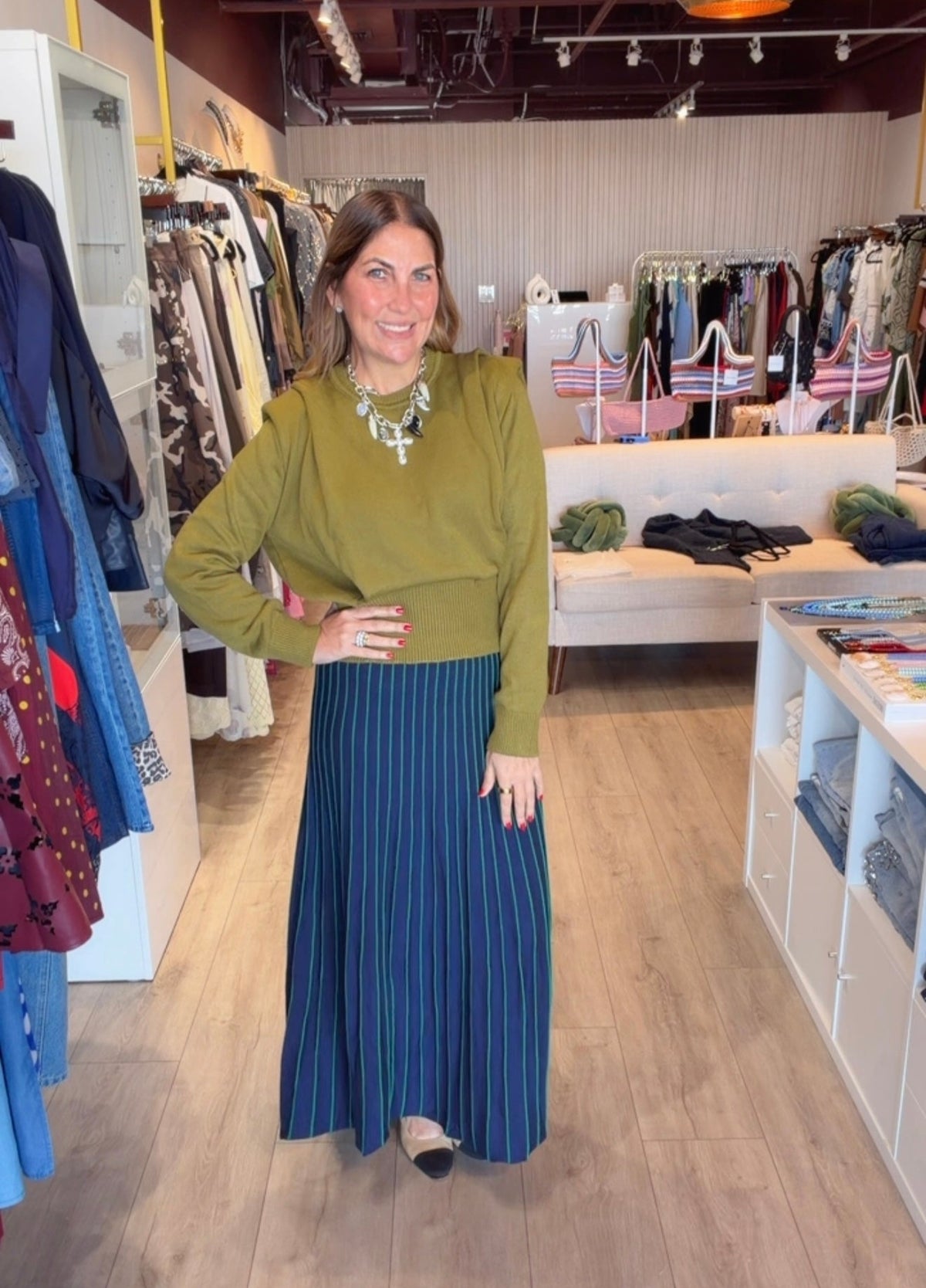 A woman modeling a sage green long-sleeve top and a navy blue pleated maxi skirt with thin white vertical stripes, standing inside a clothing boutique.