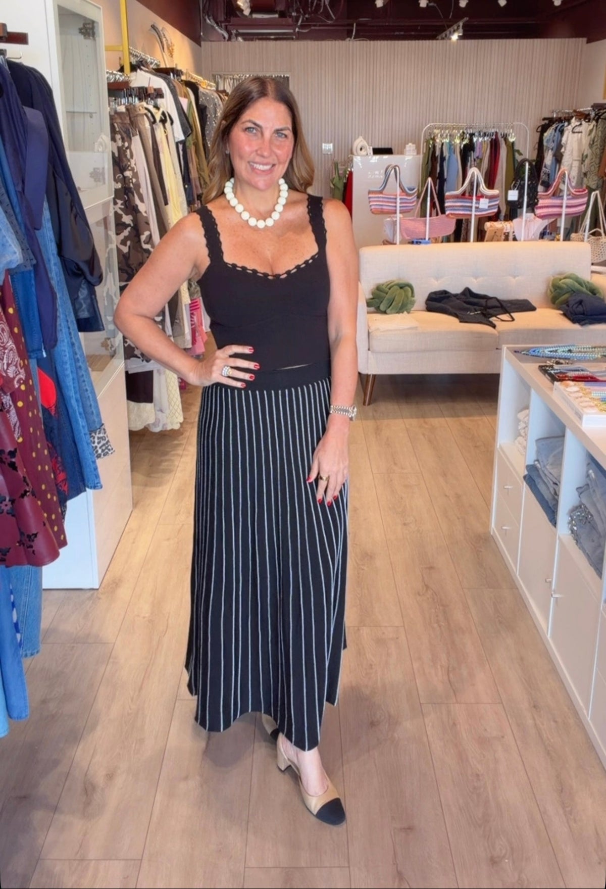 A woman modeling a black sleeveless top with gold trim and a black and white vertically striped midi skirt, standing inside a clothing boutique.