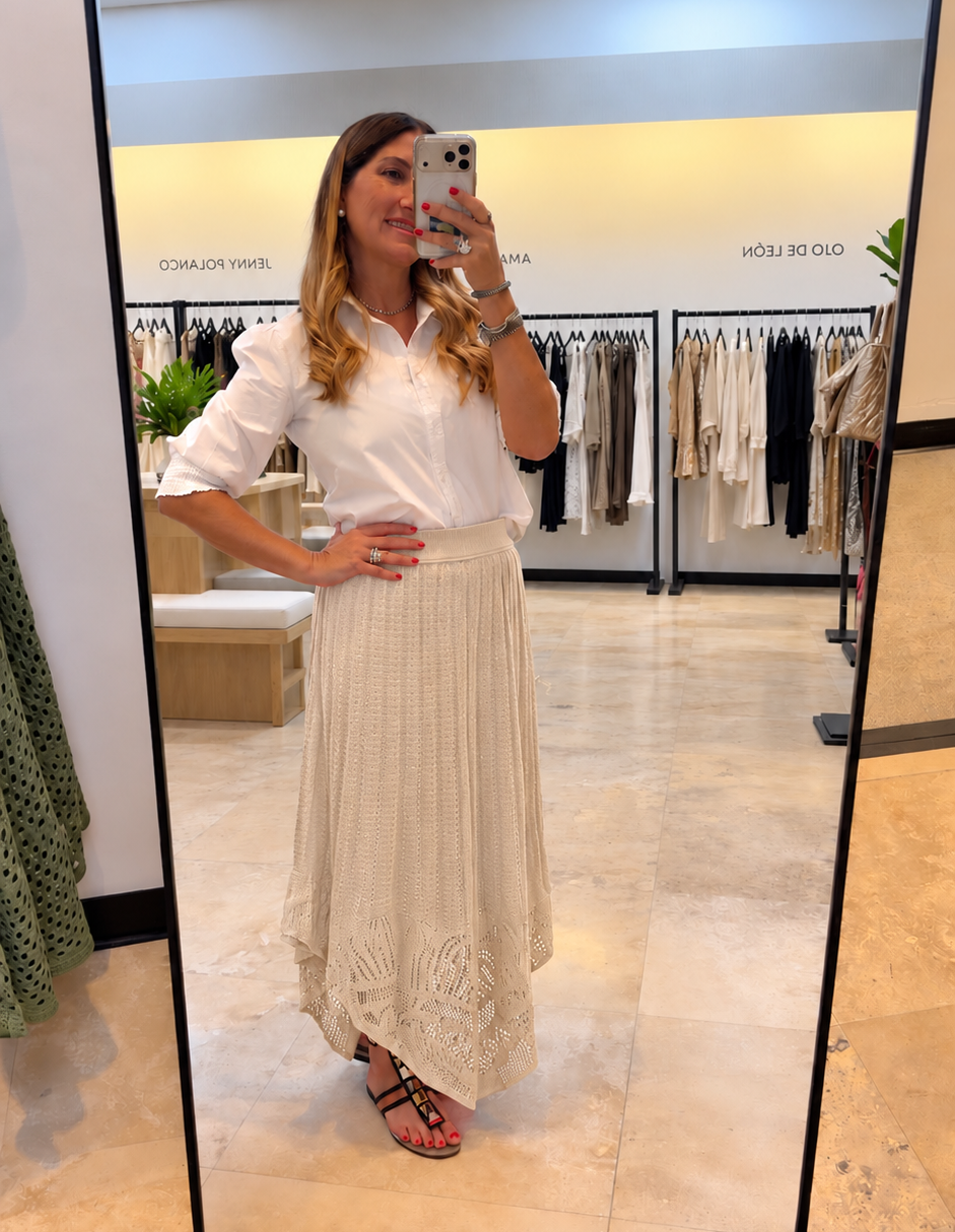 A woman takes a mirror selfie in a clothing boutique, wearing a white short-sleeve blouse with ruffled shoulders and a long cream pleated skirt with lace detailing. The background shows clothing racks and a tiled floor.