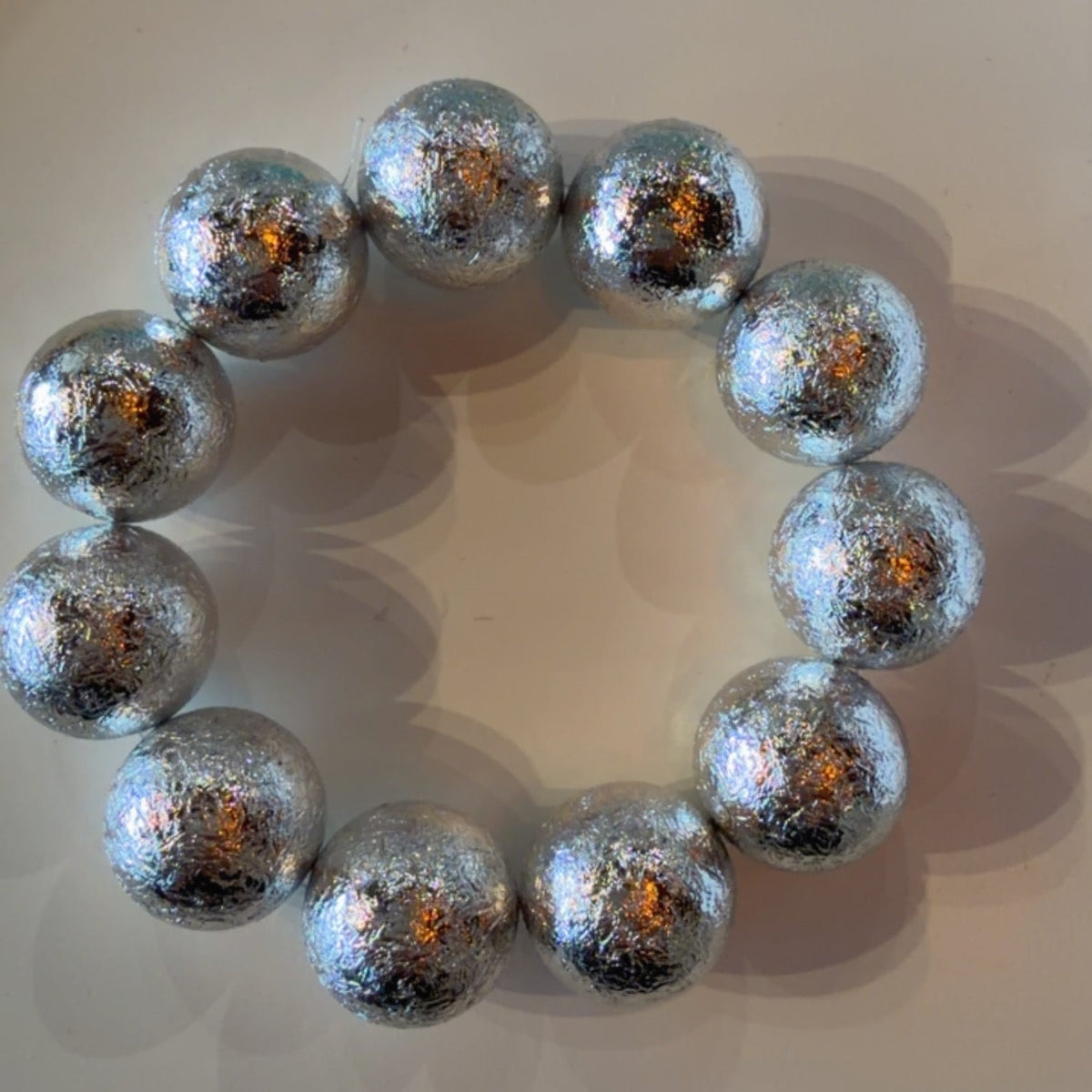 A beaded bracelet made of metallic silver beads with a textured, glittery finish, arranged in a circle on a light beige surface.