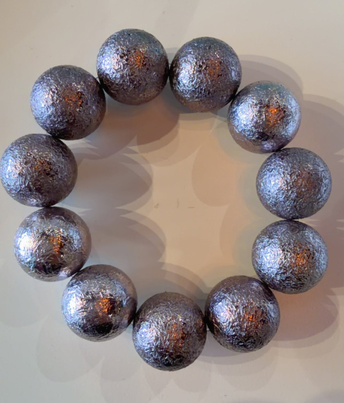 A beaded bracelet made of large metallic silver beads with a textured, glittery finish, arranged in a circle on a light beige surface.