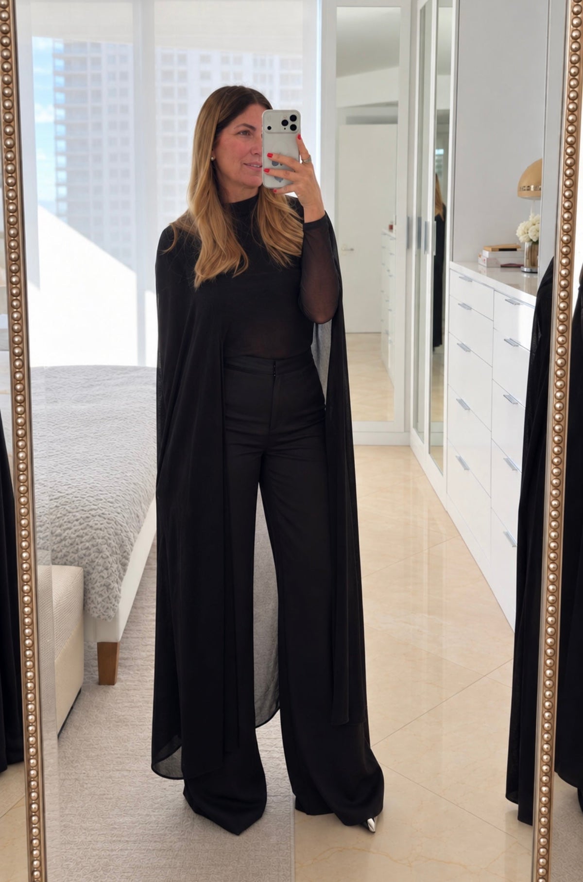 A woman takes a mirror selfie in a bright room, wearing a black long-sleeve mock-neck top with wide-leg pants and a flowing black cape-style overlay.