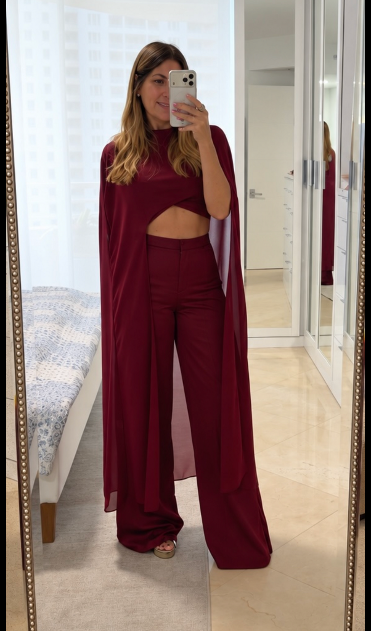 A woman takes a mirror selfie in a bedroom showing a matching burgundy two-piece outfit with a draped cape-style top and wide-leg pants.