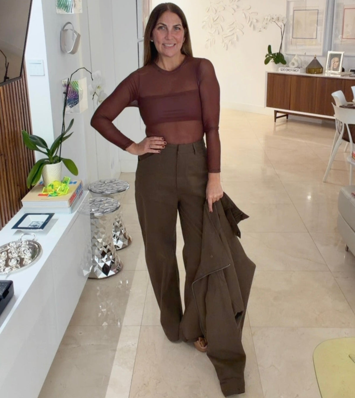 A woman stands in a modern, brightly lit home interior wearing a brown sheer long-sleeve crop top and matching brown wide-leg pants, holding a brown jacket.