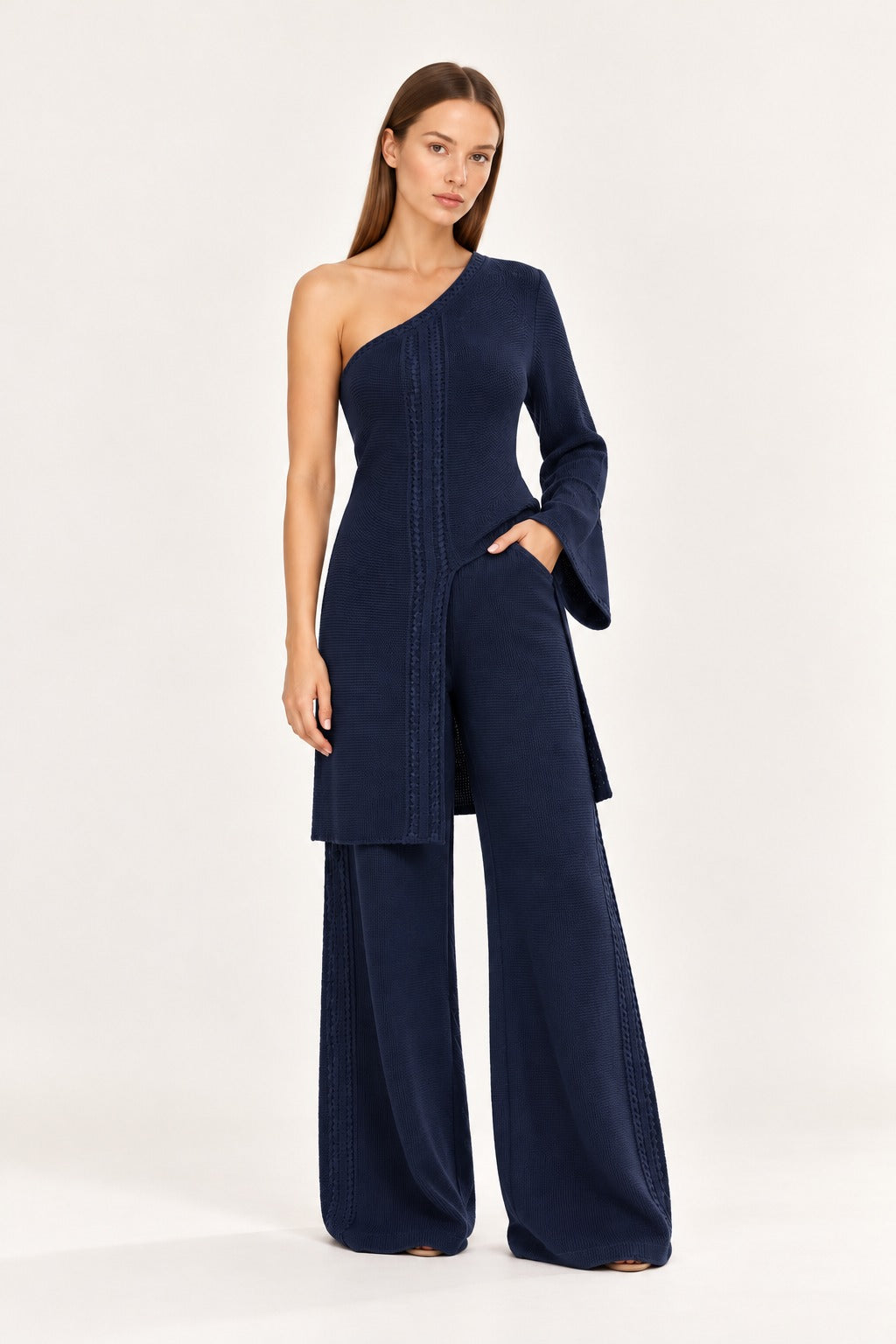 A woman modeling a navy blue one-shoulder jumpsuit with a long bell sleeve, matching wide-leg pants, and vertical seam detailing, set against a plain light background.