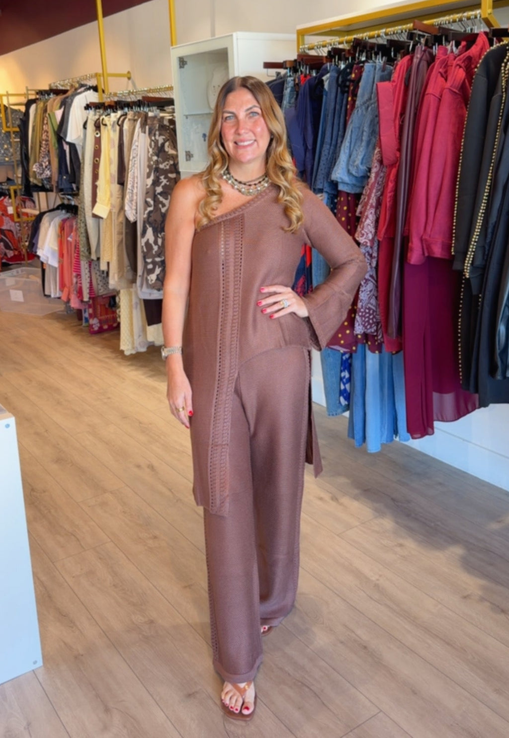 A woman modeling a brown one-shoulder jumpsuit in a clothing store, with racks of colorful garments and wooden floors visible in the background.