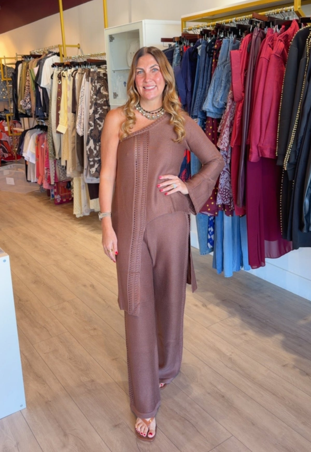 A woman modeling a brown one-shoulder jumpsuit in a clothing store, with racks of colorful garments and wooden floors visible in the background.