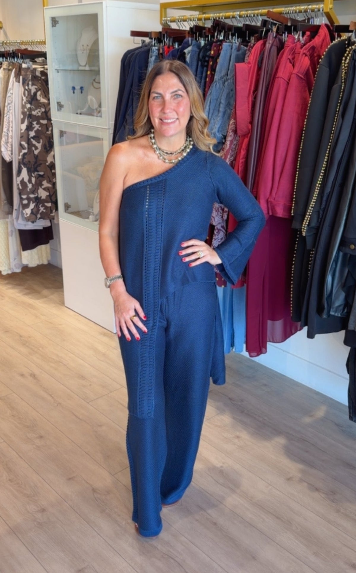A woman modeling a slate blue one-shoulder jumpsuit in a clothing boutique, standing in front of racks of garments.