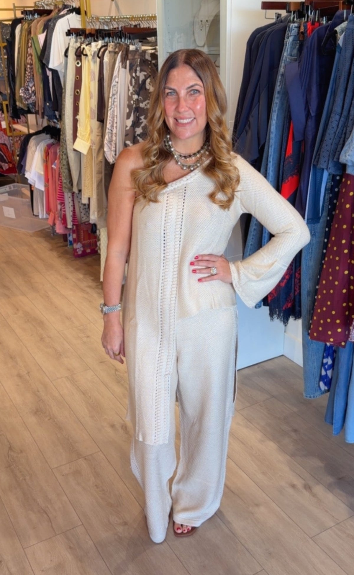 A woman modeling a cream-colored one-shoulder jumpsuit in a clothing boutique, standing in front of racks of garments on a wood floor.