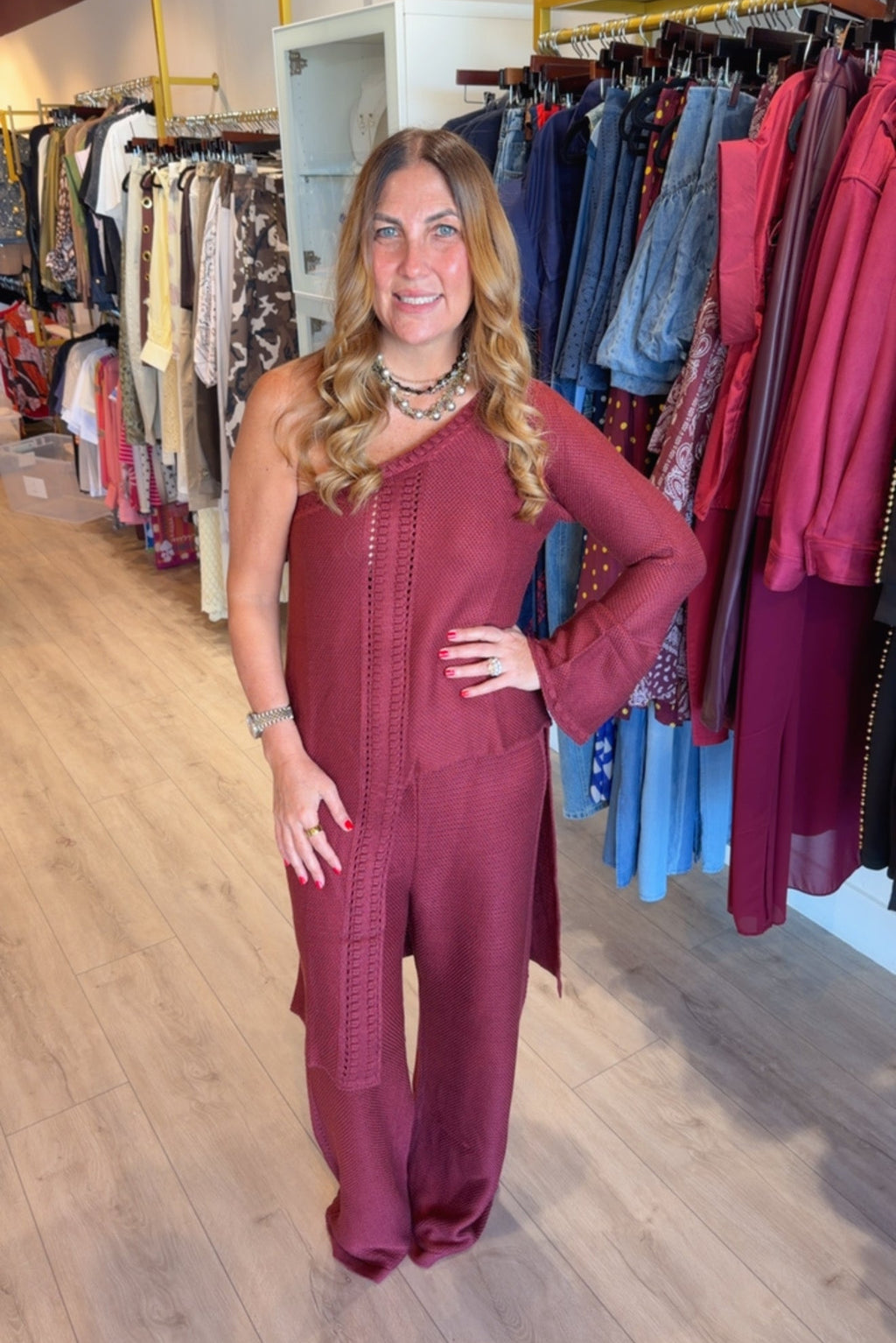 A woman modeling a rust-brown long-sleeve jumpsuit with a textured front panel, standing in a clothing boutique with racks of garments in the background.