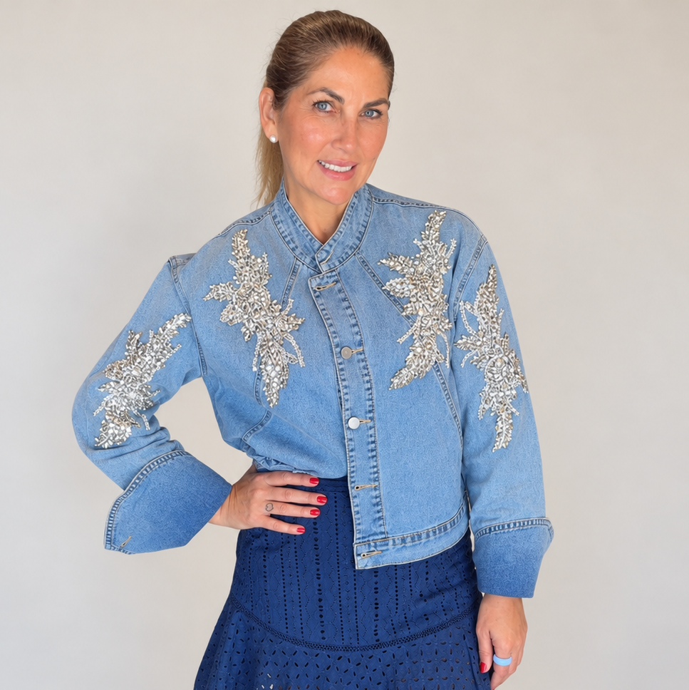 A woman modeling a light blue cropped denim jacket with silver floral embroidery and a dark blue eyelet mini skirt, paired with white sandals, standing on a light wood floor against a neutral wall.