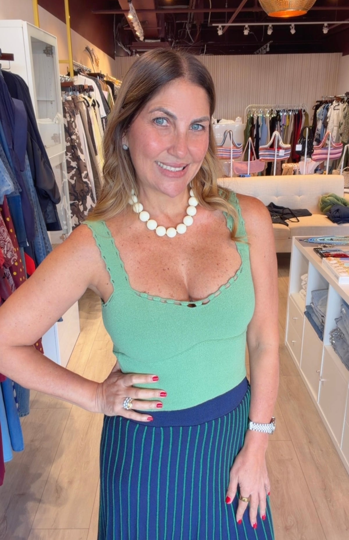 A woman posing in a clothing boutique wearing a sage green knit top with scalloped edges and a navy blue skirt with thin white vertical stripes.