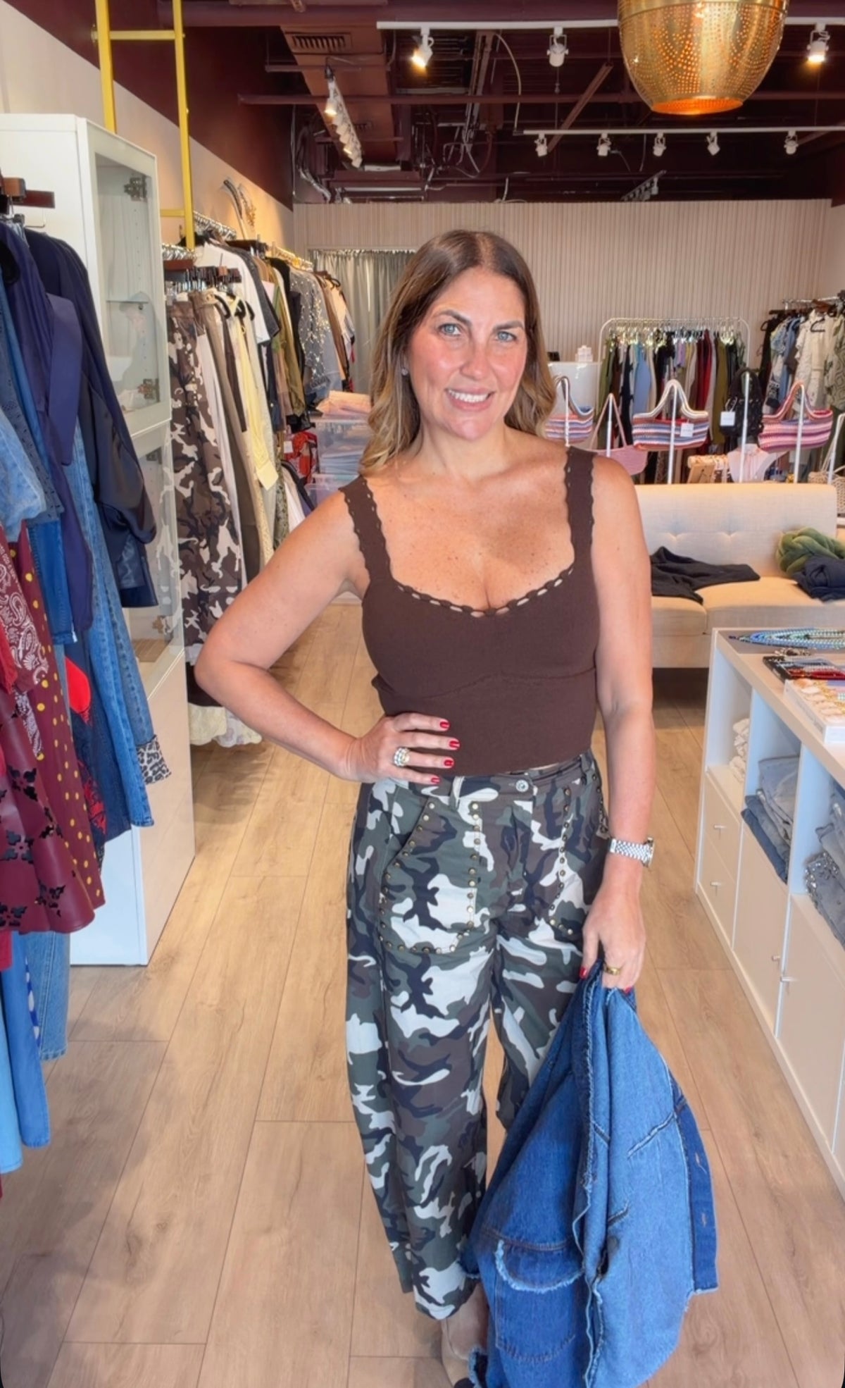 A woman posing inside a clothing boutique, wearing a brown scoop-neck tank top with lace-up straps and high-waisted camouflage pants. She is holding a blue denim jacket.